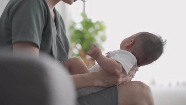 asian woman mother holding happy baby infant playing and kissing on sofa in living room at minimal style home in day time. candid authentic shot life at home. mom play with baby girl by love and care.