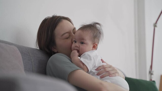 asian woman mother holding happy baby infant playing and kissing on sofa in living room at minimal style home in day time. candid authentic shot life at home. mom play with baby girl by love and care.