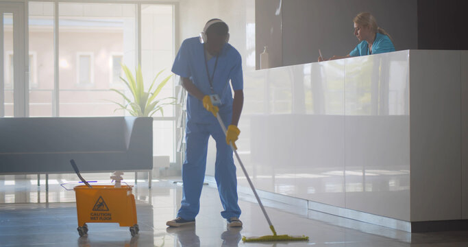 Afro-american janitor in headphones washing floor in hospital reception area