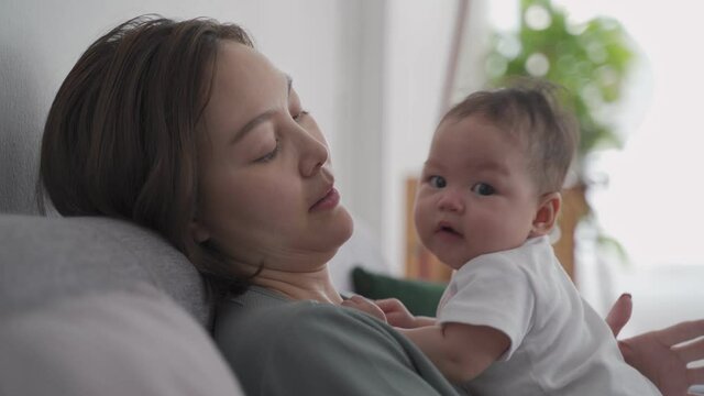 asian woman mother holding happy baby infant playing and kissing on sofa in living room at minimal style home in day time. candid authentic shot life at home. mom play with baby girl by love and care.