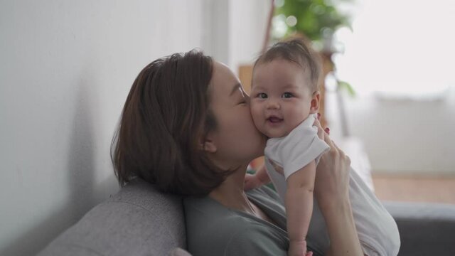 asian woman mother holding happy baby infant playing and kissing on sofa in living room at minimal style home in day time. candid authentic shot life at home. mom play with baby girl by love and care.