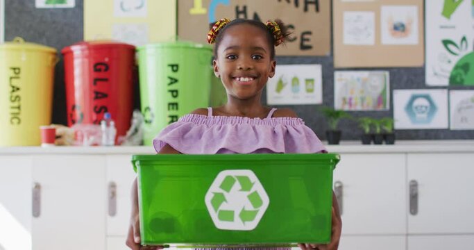 Happy african american schoolgirl standing in classroom, smiling, holding recycling bin