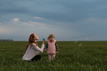 Mom plays with her daughter in the green field. A little girl watches her mother blow bubbles