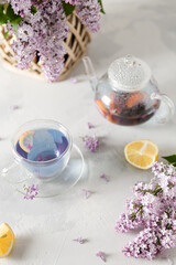 Purple tea in cup, teapot, lemons and a bouquet of blooming lilac on light background. Spring tea drinking. Greeting card, invitation design. Cafe menu, poster. Lilac tea recipe. Flat lay, copy space