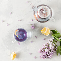 Purple tea in cup, teapot, lemons and blooming lilac on light background. Spring tea drinking. Greeting card, invitation design. Cafe menu, poster. Lilac tea recipe. Flat lay, copy space. Square