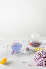 Obraz na płótnie Canvas Cup of purple tea with lemon, teapot, bouquet of blooming lilac on light background. Spring tea drinking. Greeting card, invitation design. Cafe menu, poster. Lilac tea recipe. Side view, copy space