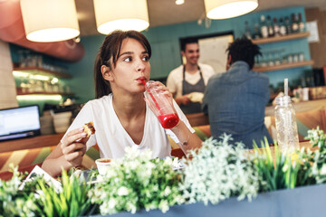 Teenage girl sitting in fast food restaurant eating burger and drinks fresh organic juice.