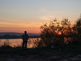 Landscape, sunset, beautiful sky. Lake shore with trees. A man in full growth looks at the other side, view from the back. Loneliness. Standing.