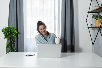 Young happy satisfied freelancer business woman taking a break from the work at home in the home office and drinking coffee from the cup chatting on the online video call with her colleague 