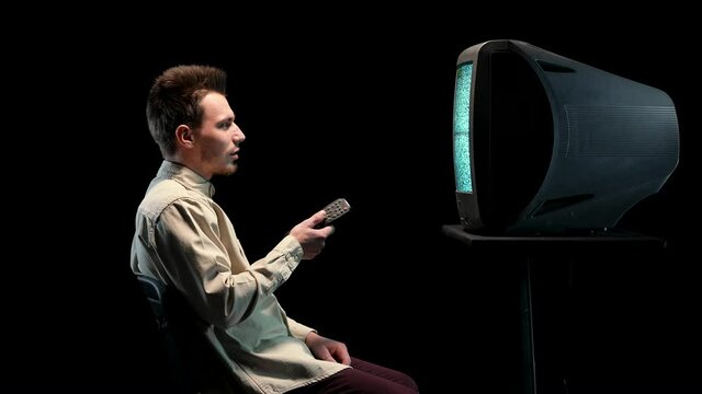 A man holds a working remote control and changes channels on an old TV, smiles, surprised in shock. Side view of a man sitting on a chair in a dark studio on a black background. Close up.