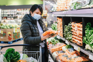 Grocery shopping. Middle age woman with short dark hair in blue protective face mask choosing buying vegetables food in supermarket and putting in shopping cart. New normal at coronavirus covid-19