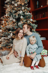Winter holidays with family. Smiling happy Caucasian mother and father with baby girl daughter by decorated Christmas tree at home. Happy family celebrating Christmas or New Year together