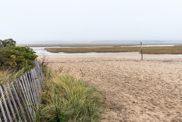 View of coastal marsh on a foggy autumn morning. A weathered fence at the foot of grassy sand dunes is in in foreground. Sandwich, Cape Cod, MA, USA.