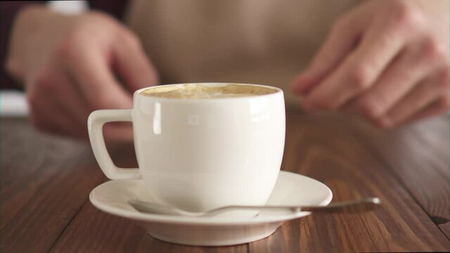 A male barista in a beige apron serving a cup of freshly made cappuccino. Male hands move a white cup of Americano coffee on a wooden table. Coffee on the table to serve guests. Coffee bar, small busi