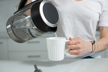 A girl in a white T-shirt pours clean water from the kettle into a white mug. Healthy lifestyle. High quality photo