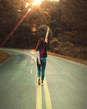 Mulher andando no meio da rua em uma estrada com curva e árvores a beira do caminho ao pôr do sol. A mulher usa calça jeans e camiseta preta e tem uma das mãos erguidas.