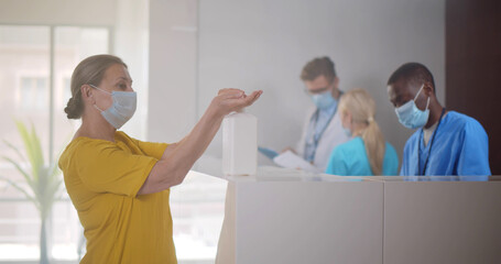 Senior patient in safety mask disinfecting hands with antibacterial gel at reception desk in...