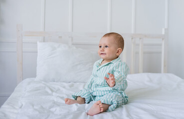 Fototapeta premium baby boy in striped pajamas sits on the white bedding on the bed and looks away. Space for text