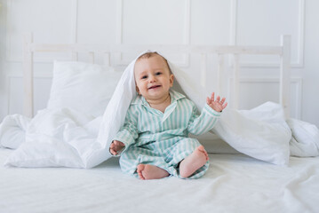 happy baby boy in striped pajamas sits under a white cotton blanket on the bed. Healthy sleep