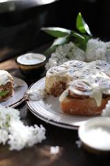Cinnamon rolls covered with butter cream on a table with white lilacs. Breakfast with rolls and milk.