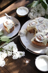 Cinnamon rolls covered with butter cream on a table with white lilacs. Breakfast with rolls and milk.