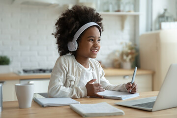 Smiling small African American girl child in headphones study online on computer at home. Happy little biracial kid in earphones write in notebook have webcam lesson on laptop. Education concept.