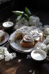 Cinnamon rolls covered with butter cream on a table with white lilacs. Breakfast with rolls and milk.