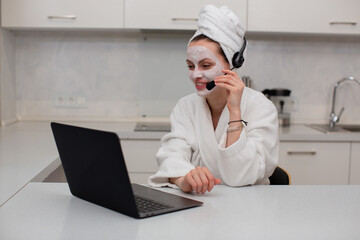 A young girl with a moisturizing mask on her face in a white robe talking about the headset working behind a laptop, sitting in the kitchen. High quality photo