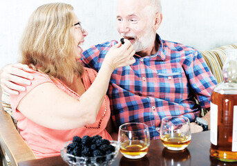 Cheerful senior couple tasting whiskey at home. Woman treats a man with a berry