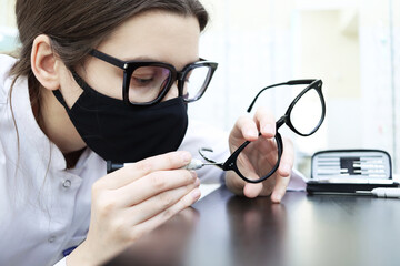 Repair of eyeglasses. Optical salon and workshop. A young woman in a medical gown and a protective mask fixes medical glasses. Tightening the screw into the temple of the spectacle frame