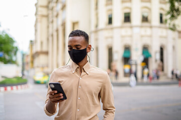 African businessman outdoors in city wearing face mask and using mobile phone