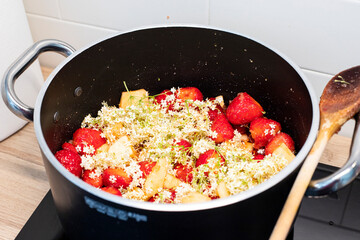 Strawberries and elderflowers jam cooking in black pot with wooden spoon. Spring foraging preserve recipe inspiration. Close-up, side view.