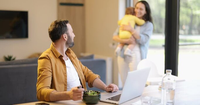 Young father works on a laptop during a lunch time at home with his wife and one year son. Home family routine and work from home concept