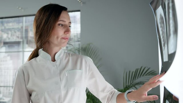 Young Female Doctor In White Coat. She Stands Near An Xray of The Lungs And Tells Diagnosis.