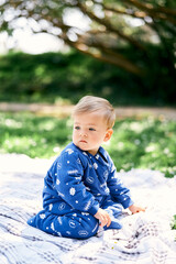 Small child in a blue overalls sits on his knees with his head turned on a checkered blanket on a green lawn