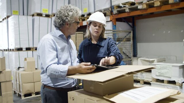 Serious bearded boss talking with female worker using tablet. Caucasian man in eyeglasses and woman in helmet having conversation about delivery of goods. Manufacture, industrial occupation concept.