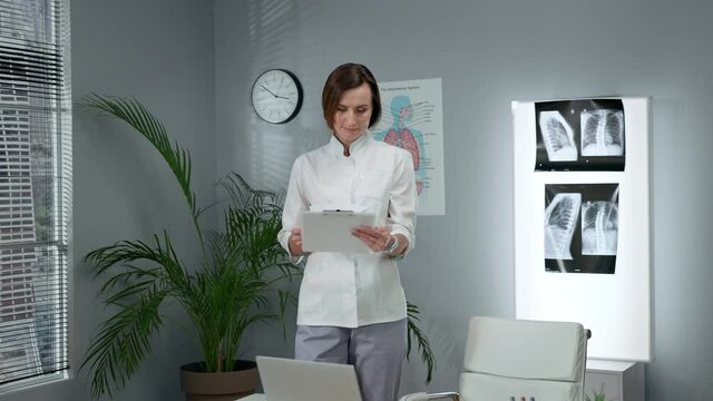 Young Female Doctor In White Coat. Woman Stands In The Middle Of The Office And Flips Through The Sheets With Tests.