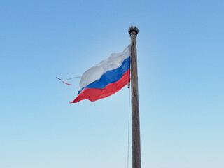 The white-blue-red flag of the Russian Federation flutters in the wind on an old wooden flagpole against a bright blue sky without clouds. The concept of news about Russia.