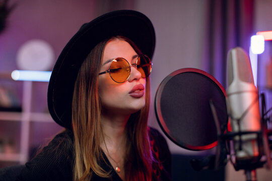 Portrait of charming woman in stylish black hat and sunglasses singing in professional microphone indoors. Female musician recording new track at modern studio.