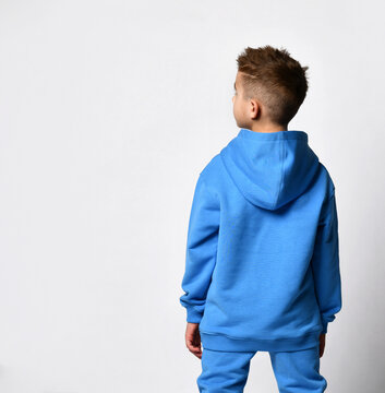 Schoolboy boy in sportswear stands with his back to the camera on a white background. Back view of a child in a blue sports hoodie and pants standing near a free space for text. Fashion and sports.