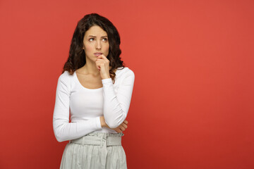 Obraz premium Portrait of serious doubtful woman looking up thinking pensive and confused. Young curly girl in white outfit isolated on red background. Concerned upset female in 30s bite lips in doubts and thoughts