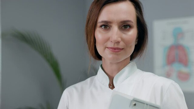 Portrait Of White Female Doctor In White Coat. Woman Standing In The Office Holding Notebook.