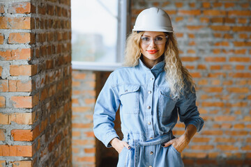 Attractive female construction worker in hardhat. Confident young specialist in checkered blue shirt in jeans standing in empty room. Interior design and renovation service.