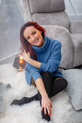 beautiful young girl with red hair and a blue sweater sits near a gray armchair in the living room by the panoramic window