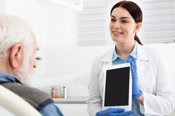 Obraz premium cheerful dentist showing tablet to senior patient in dental clinic.