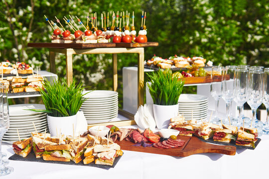 open-air buffet table, glasses and sandwiches on skewers before the start of the holiday against the background of flowering trees in the garden