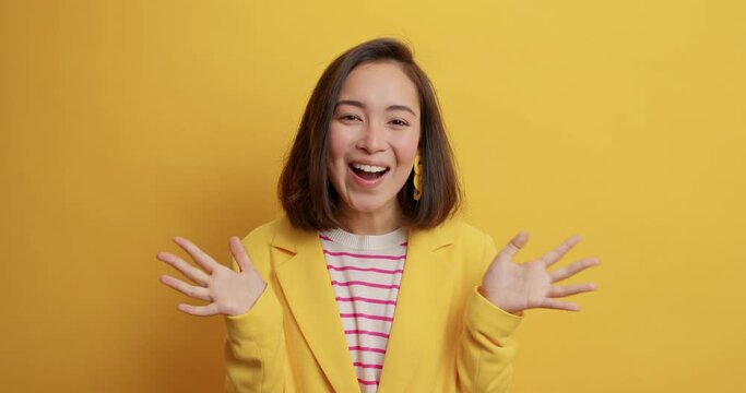 Surprised cheerful Asian woman clasps hands hears awesome news smiles happily exclaims emotionally glad to hear about own success and achievements dressed elegantly isolated on yellow studio wall