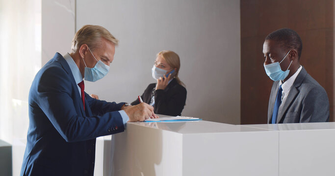 Businessman in safety mask signing document from receptionist in office