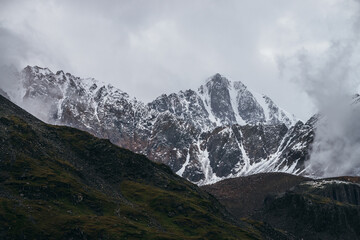 Atmospheric alpine landscape with great mountain peaked top with snow in low clouds. Dramatic mountain scenery with sharp pinnacle in overcast weather. Awesome view to snowy pointy peak in cloudy sky.