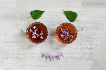 The concept of good morning and good mood. Two cups of tea with lilac petals, two green leaves and flowers laid out in a cheerful face on a gray background, top view.